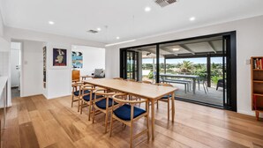 A spacious room with a wooden dining table and chairs, a large sliding glass door leading to an outdoor patio, and a bookshelf. - Coastal Retreat—Treetop Outlooks by Goolwa Beach (Goolwa South)