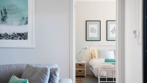 A modern bedroom with a white bed, bedside tables, and a nightstand. There are framed pictures on the wall and a window with a view of trees.