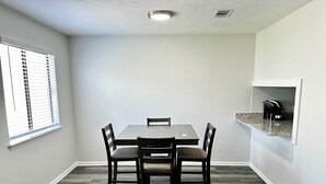 A dining area with a rectangular table and four chairs, a window with blinds, and a built-in kitchen shelf with a coffee maker. - Stadium Stay 2 Bedroom (Tallahassee)