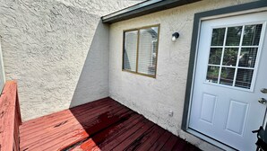 A corner of a building with a white textured wall, a red wooden deck, a white door with a window, and a small wall-mounted light. - Stadium Stay 2 Bedroom (Tallahassee)