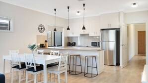 A modern kitchen with white cabinetry, a stainless steel refrigerator, and a dining area with white chairs and a table. - Brooking’s Perch - Riverside Stay near Cafés & Wharf (Goolwa)