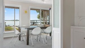 Een moderne eethoek met een grote tafel en vier stoelen, grote ramen met uitzicht op een wateroppervlak, en een plafondventilator.