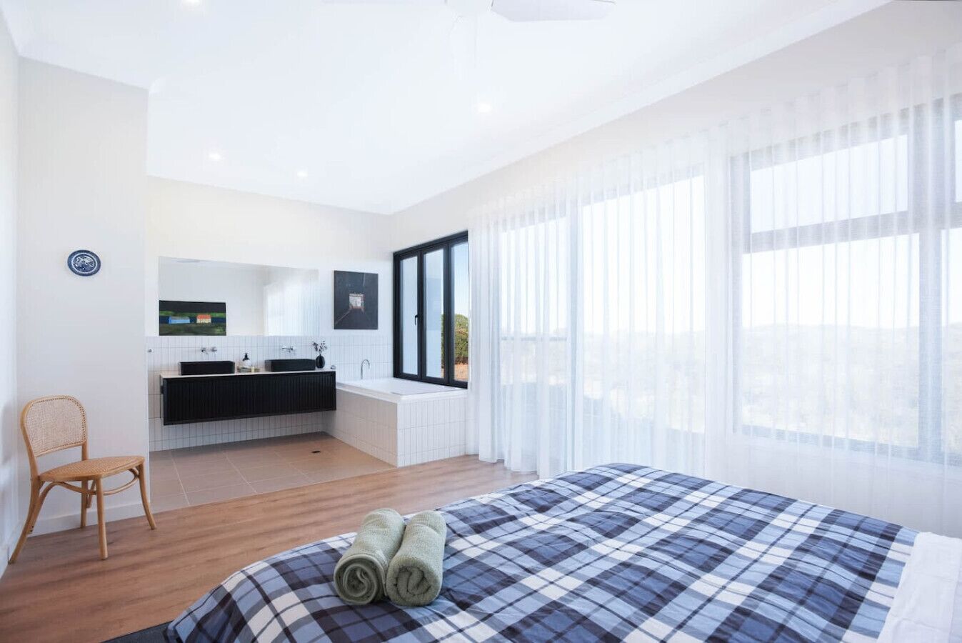A modern hotel room with a large bed, a sitting area with a chair, a bathroom with a bathtub, and a view through large windows.