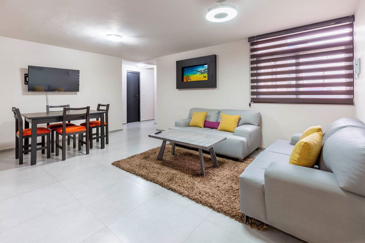 A modern living room with a dining area, a flat-screen TV mounted on the wall, a sofa with cushions, and a large window with blinds.