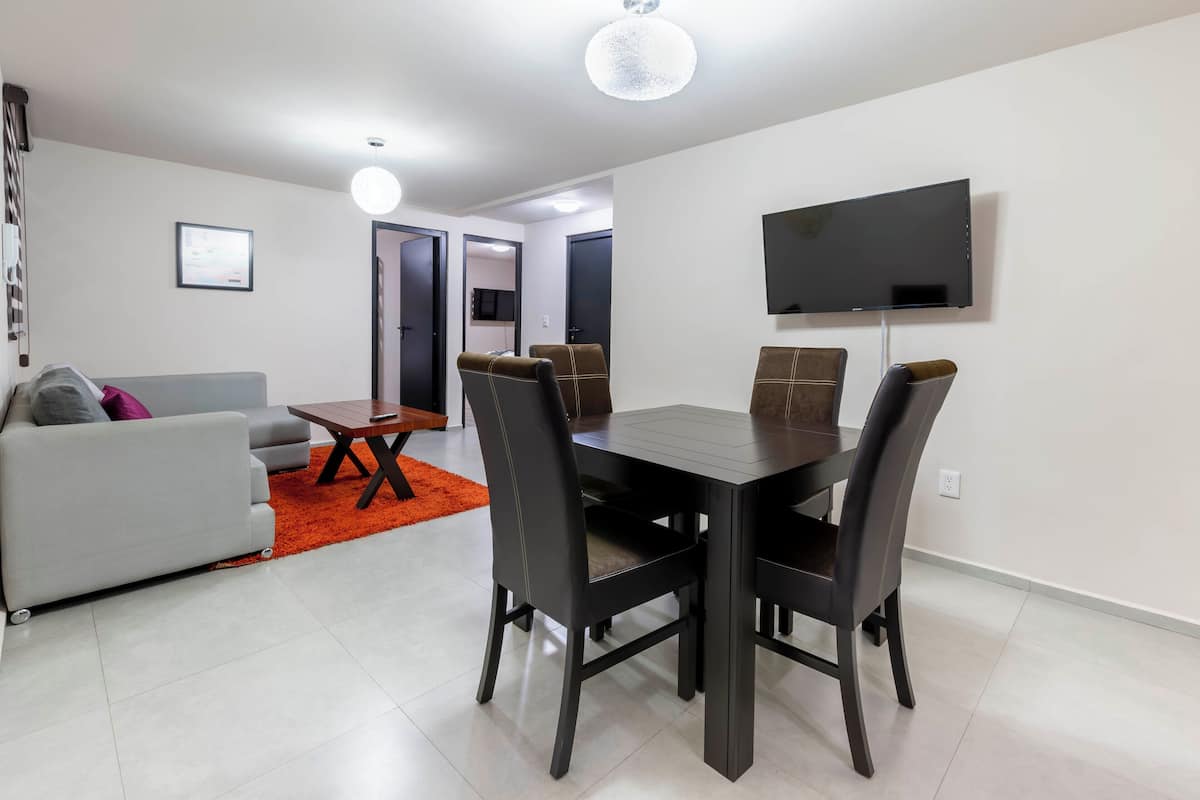 A modern living room with a grey sofa, a dining table with four chairs, a flat-screen TV mounted on the wall, and a bright orange rug.