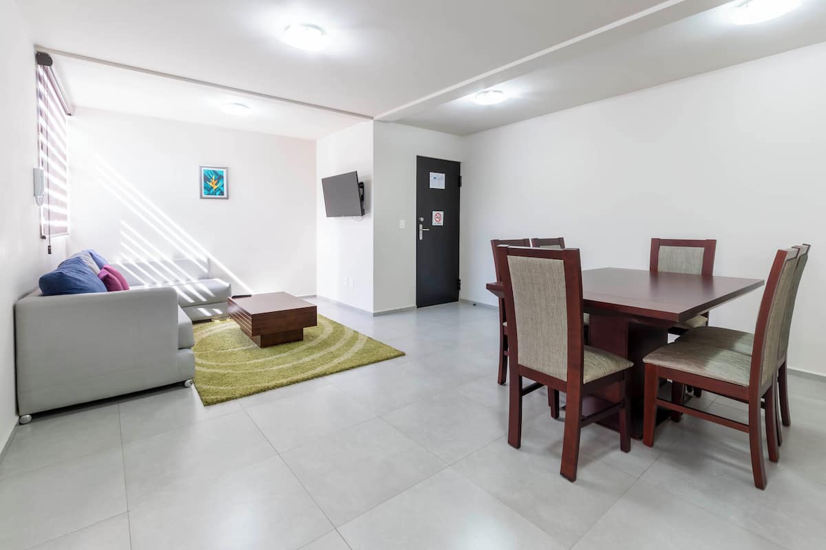 A modern living room with a sofa, a dining table with chairs, a staircase, and a door with a sign.