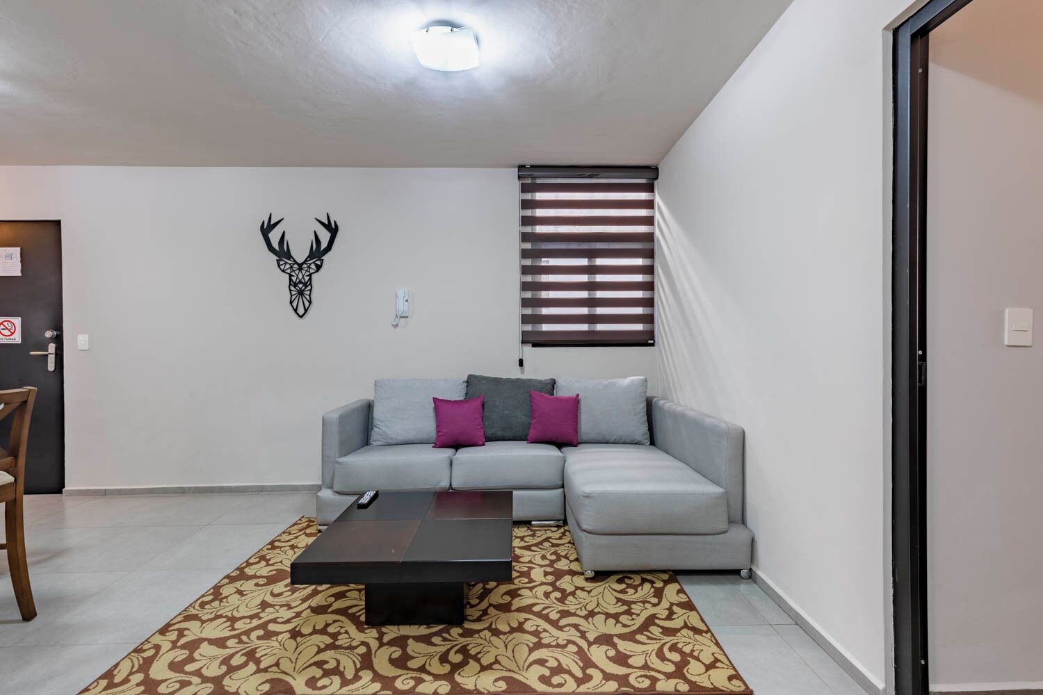 A living room with a grey sofa, a patterned rug, a wall-mounted deer head decoration, and a window with blinds.