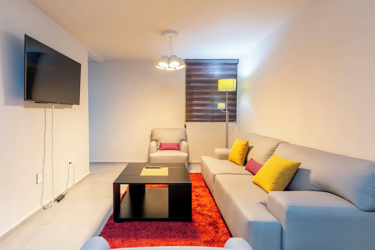 A modern living room with a flat-screen TV, a sofa with yellow and pink cushions, a red carpet, and a ceiling light fixture.