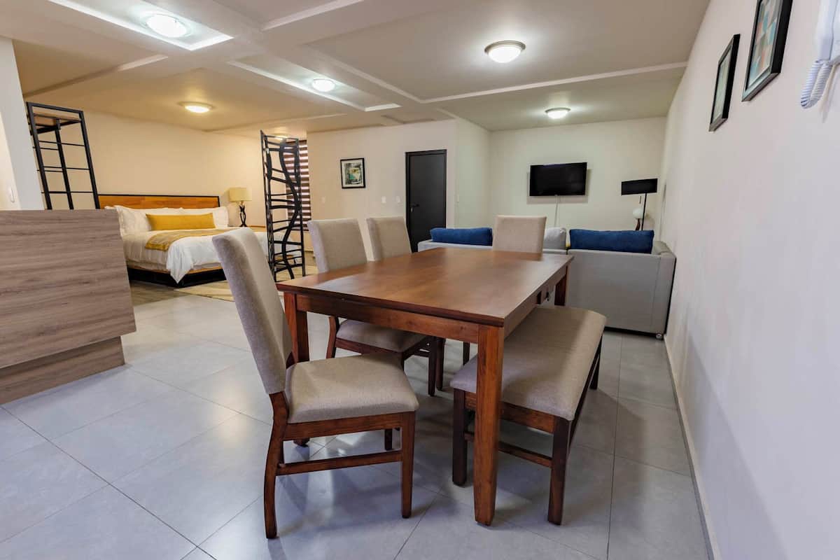 A modern living room with a wooden dining table and chairs, a sofa, a TV, and a bed with a ladder.