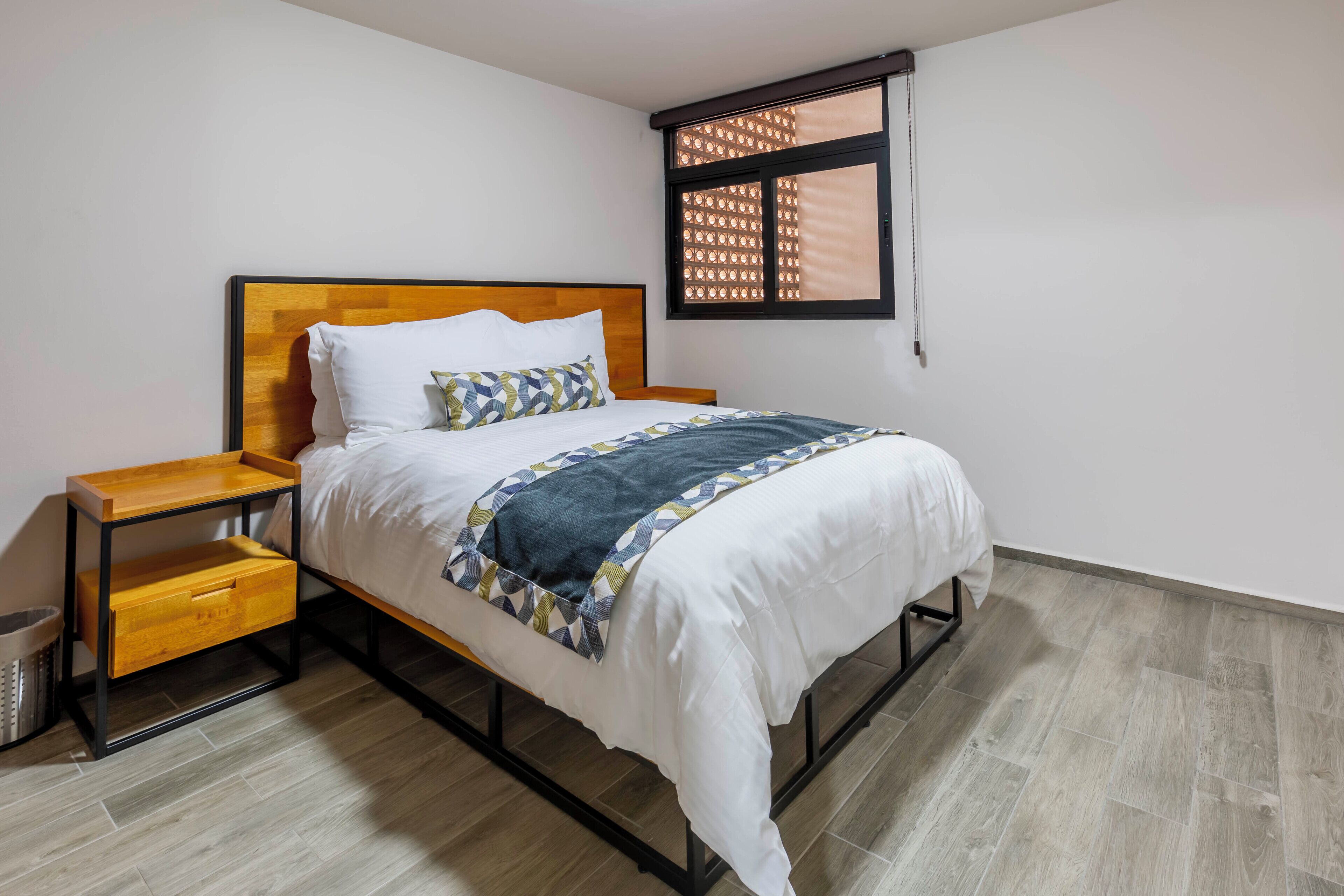 Una cama bien tendida con cabecera de madera, una mesita de noche con cajones y una ventana con persiana estampada.
