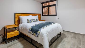 Una cama bien tendida con cabecera de madera, una mesita de noche con cajones y una ventana con persiana estampada.