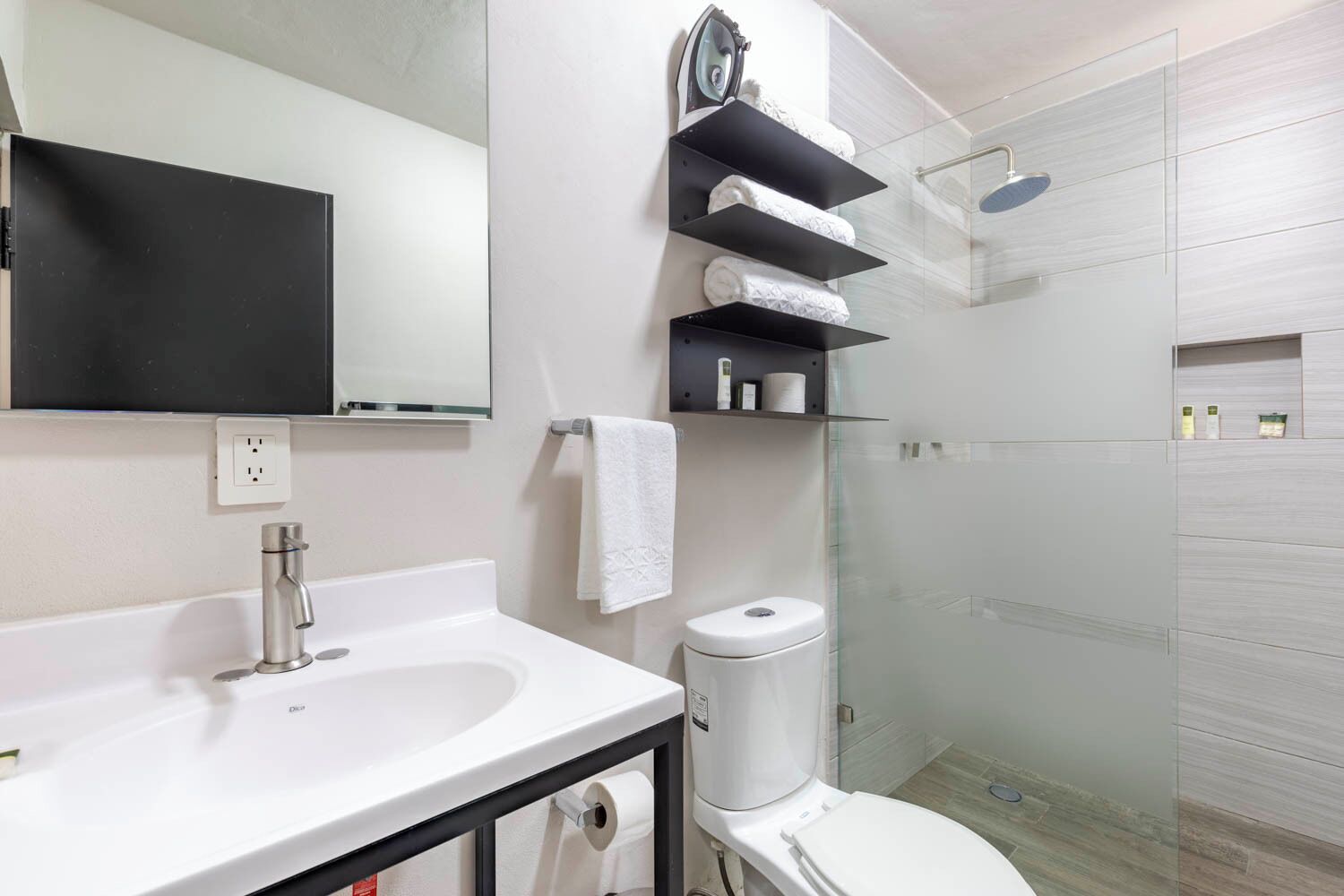 A modern bathroom with a white sink, a toilet, a glass shower enclosure, and black shelves with towels.