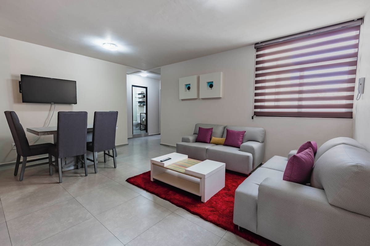 A modern living room with a dining area, a flat-screen TV mounted on the wall, a grey sofa with cushions, a red rug, and a white coffee table.