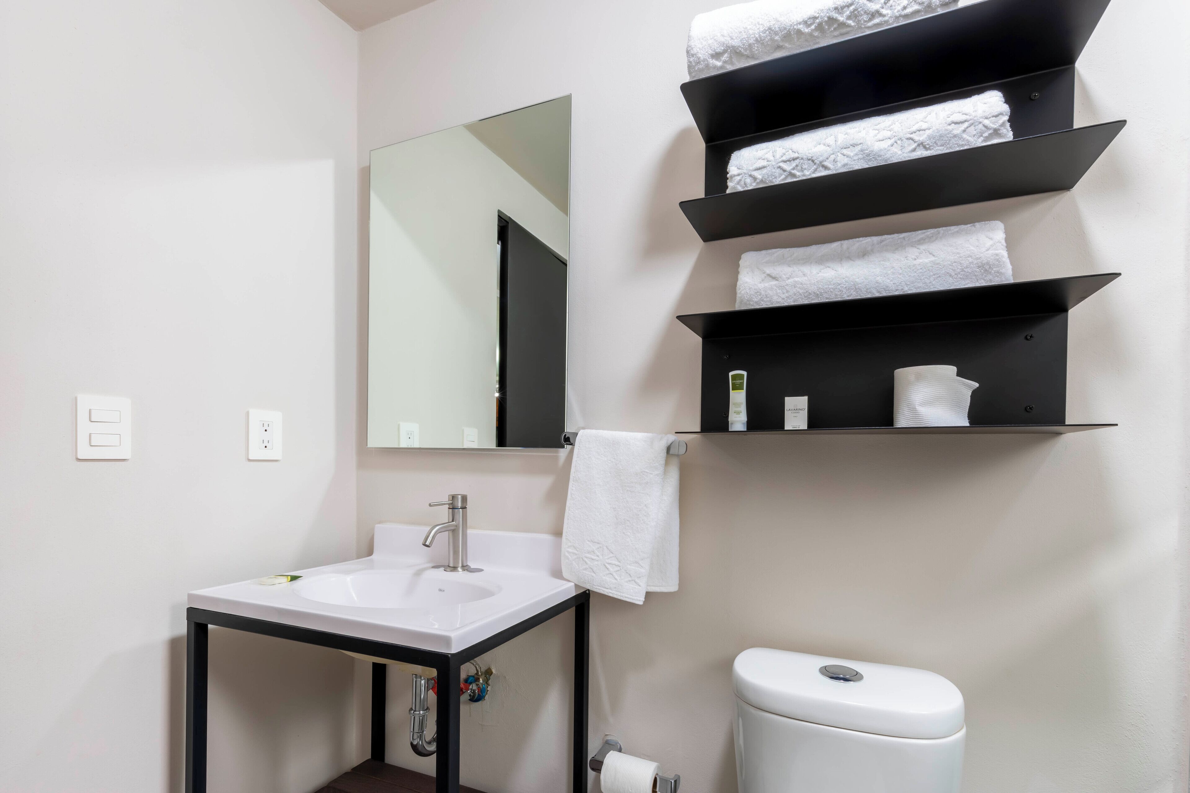 A bathroom with a white sink, a mirror, a toilet, and black shelves holding towels.