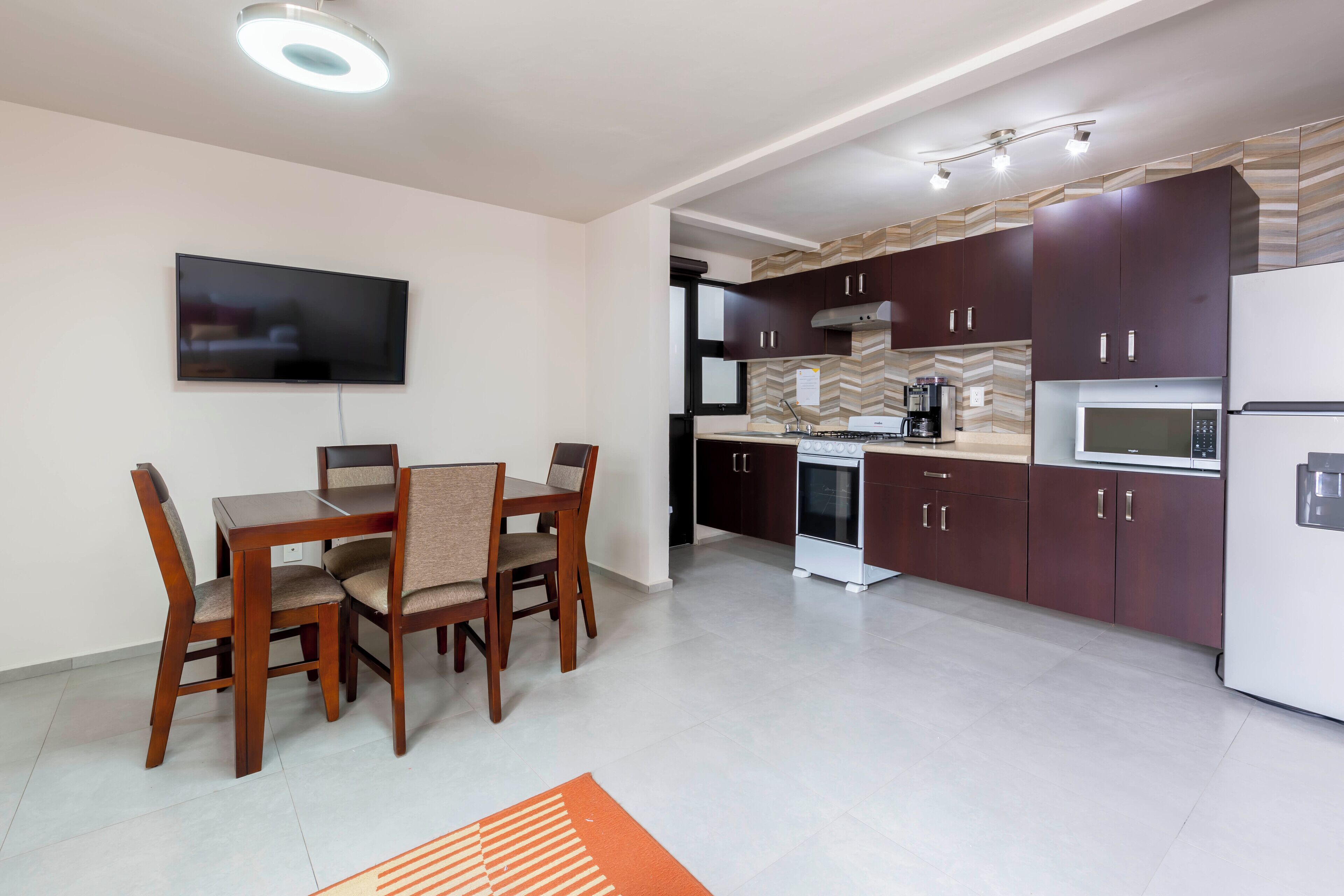 Une cuisine moderne avec des meubles en bois foncé, un réfrigérateur blanc et un four à micro-ondes. On y trouve une table à manger avec des chaises et un téléviseur à écran plat fixé au mur.