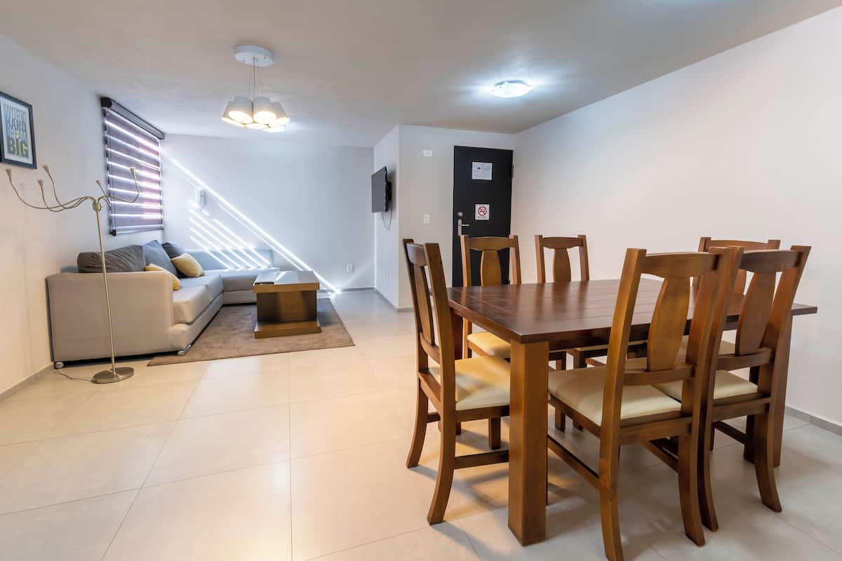 A modern living room with a sofa, a dining table with chairs, a TV mounted on the wall, and a staircase leading to another floor.