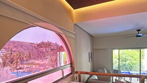 A room with a large curved window offering a view of a hillside with buildings and palm trees. There is a wooden bench and a table with two bowls. - Luxury beachfront condo with private pool & fast internet (Bahías de Huatulco)