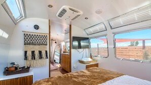 A compact, well-lit bedroom with a bed, a wall-mounted TV, and a window with a view of the outdoors.
