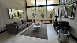Een moderne woonkamer met een grijze bank, een glazen salontafel en een groot raam met potplanten.