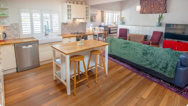 A modern kitchen with white cabinets, a wooden countertop, and a dishwasher. There is a dining area with a table and two stools. A red armchair and a blue sofa are visible in the living room area. - Bylund Cottage – Historic Charm in the Heart of Albany (MIRA MAR)