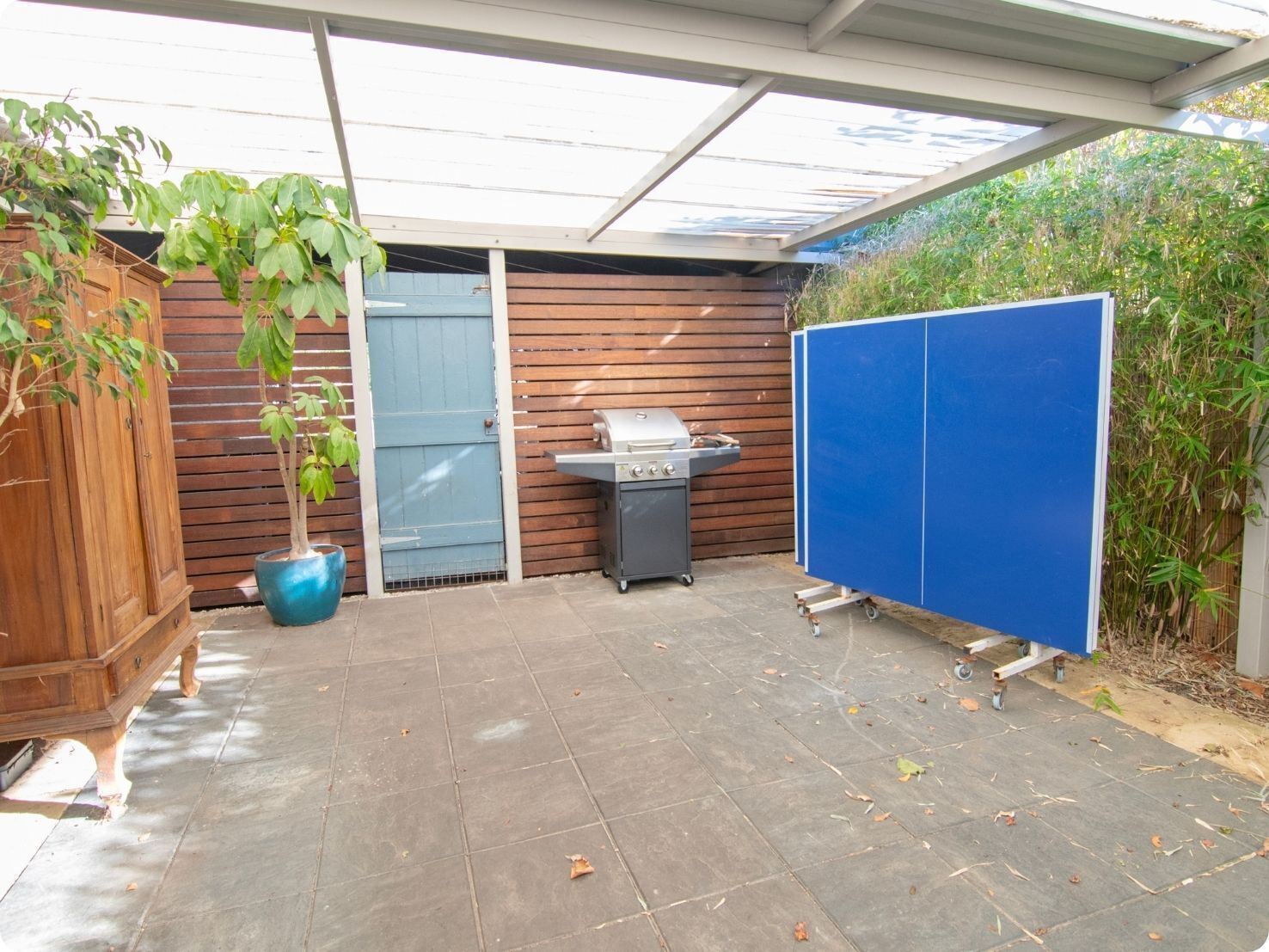 Ein überdachter Patio-Bereich mit einem Grill, einem Blumentopf und einer blauen Trennwand.