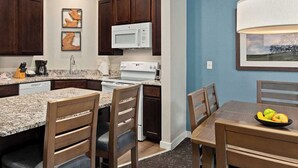 Une cuisine compacte équipée d’un réfrigérateur, d’un four à micro-ondes, d’une cuisinière et d’un plan de travail en granit. On y trouve des chaises en bois et une table à manger avec un panier de fruits.