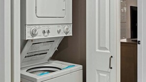 Un appareil compact combinant lave-linge et sèche-linge, installé dans un couloir étroit, avec une poignée de porte et une petite étagère au-dessus du sèche-linge.