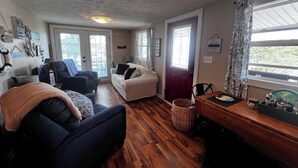 A cozy living room with a sofa, armchair, and a dining table. There are two windows with curtains, a door, and a ceiling light. - Waterfront 2BR Cottage: Fire Pit, Grill & Dock -Boat Slip Available additional (Monticello)