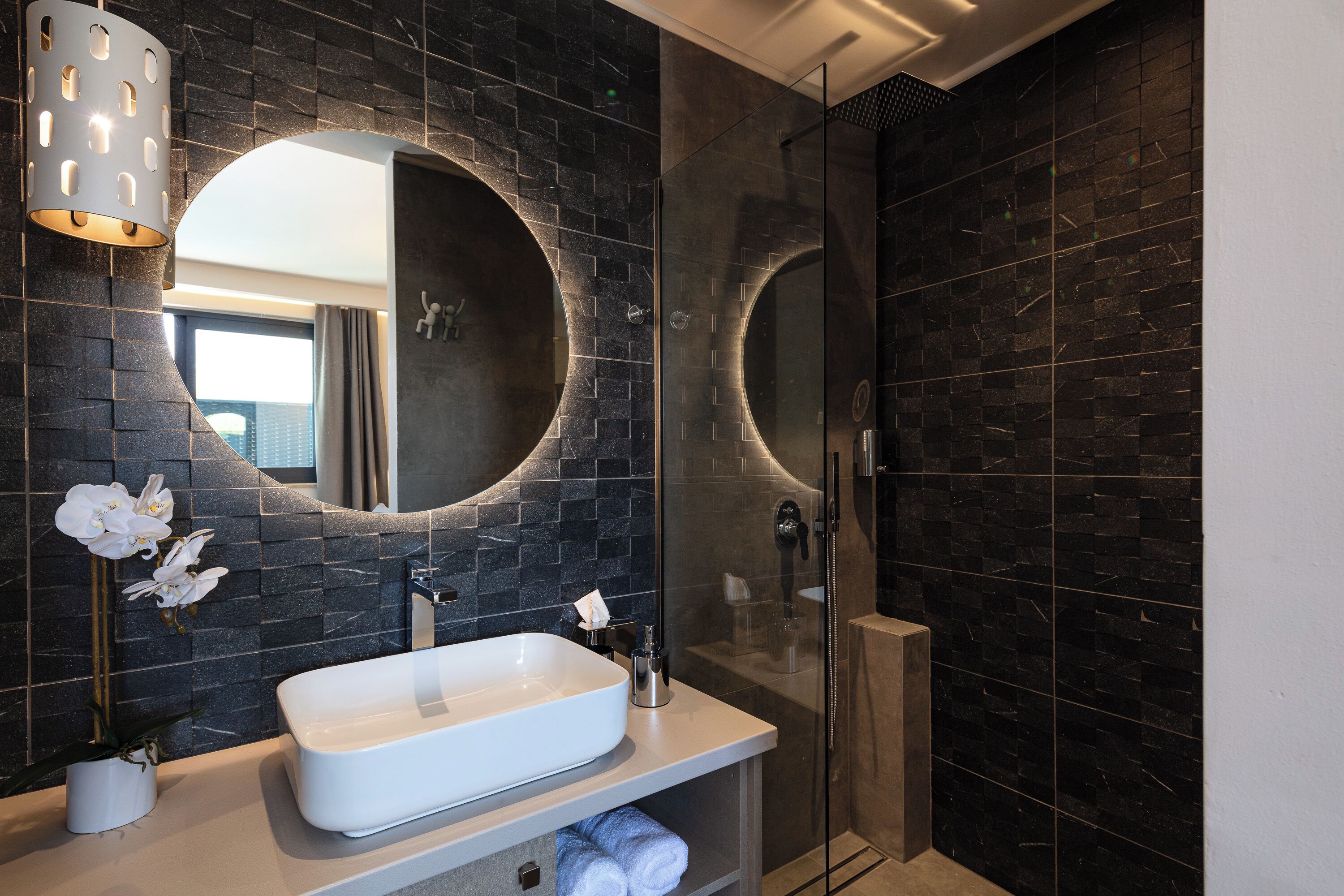 A modern bathroom with a large oval mirror, a white sink, and a glass-enclosed shower.