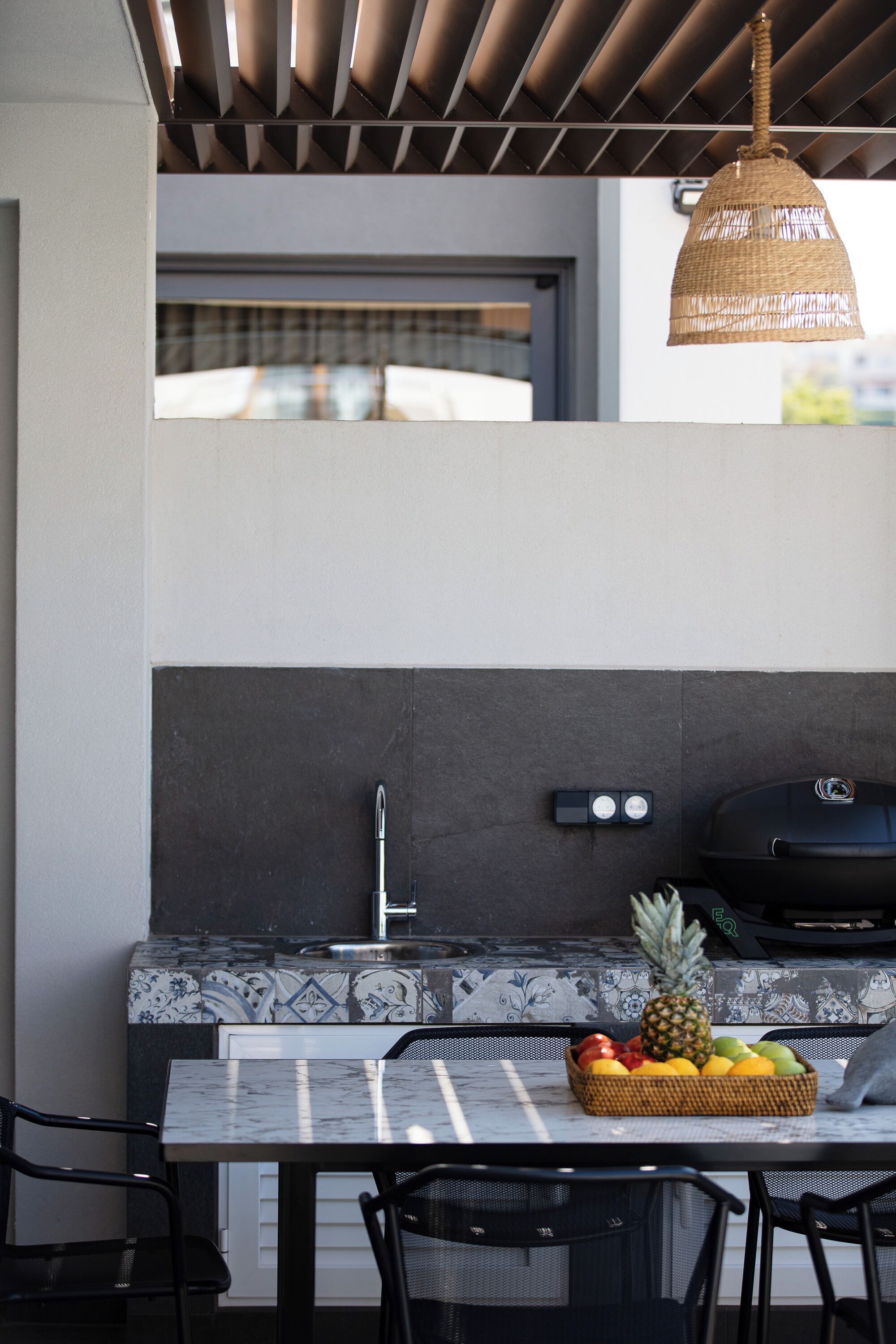 An outdoor kitchen with a tiled backsplash, a hanging light fixture, a grill, and a fruit basket on the table.