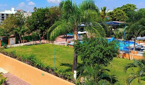 Zona de piscina con palmeras, césped y un edificio al fondo.