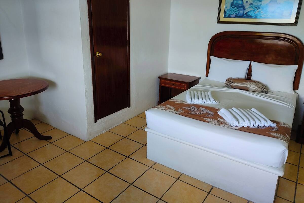 Una camera d'albergo con un letto, un tavolino in legno, un comodino e una porta.