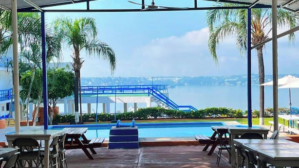 A covered outdoor area with a pool, tables, and chairs, overlooking a body of water and a bridge.