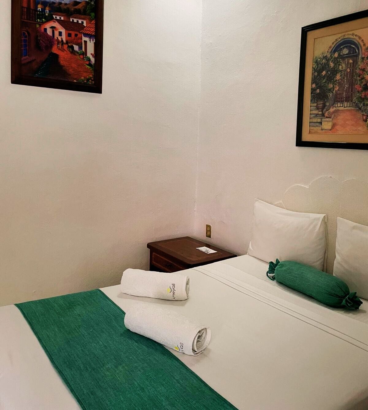 A hotel room with a bed, green bedspread, white pillows, a nightstand, and two paintings on the walls.