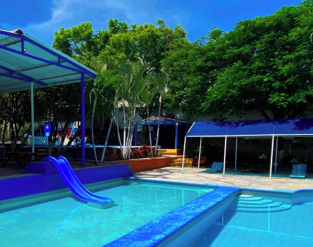 Piscina con tobogán, rodeada de árboles y zonas cubiertas.