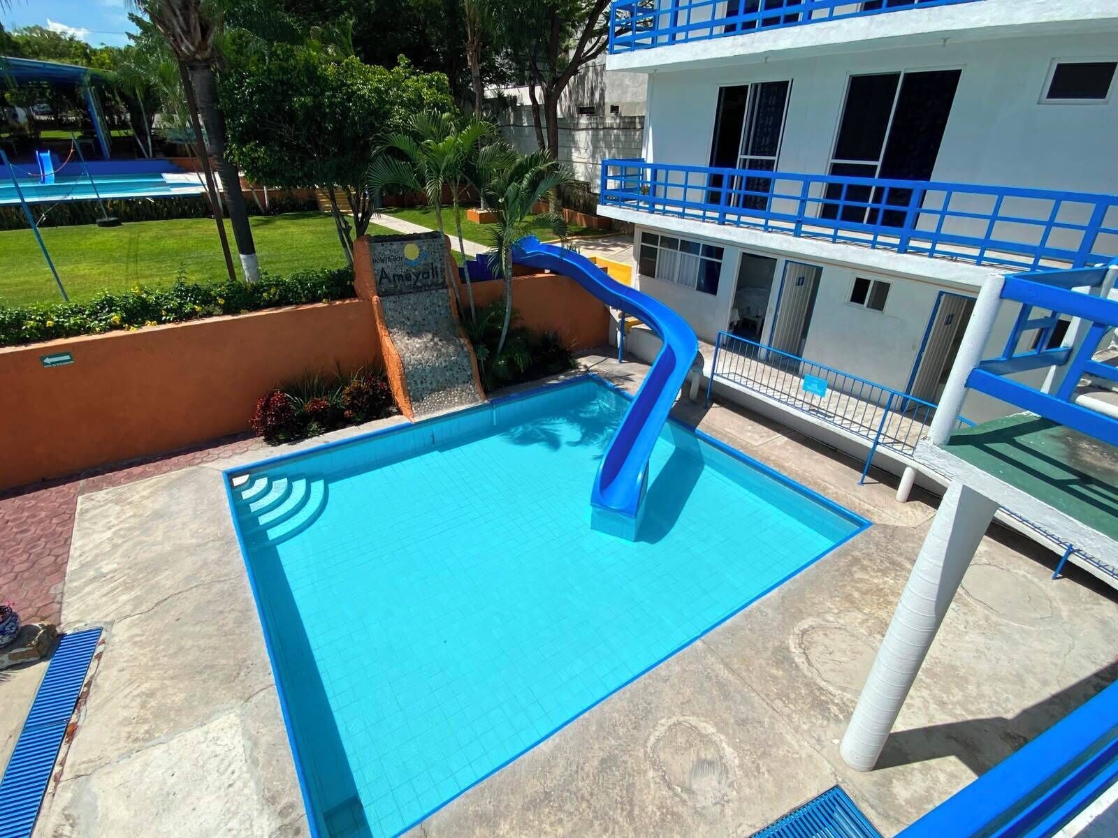 A swimming pool with a blue slide, surrounded by a white building with blue railings, and a green lawn with palm trees.
