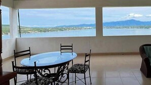 A room with a dining table and chairs, a sofa, and large windows with a view of a lake and mountains.