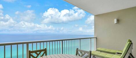 A balcony with a wooden table and two chairs, a lounge chair, and a view of the ocean.
