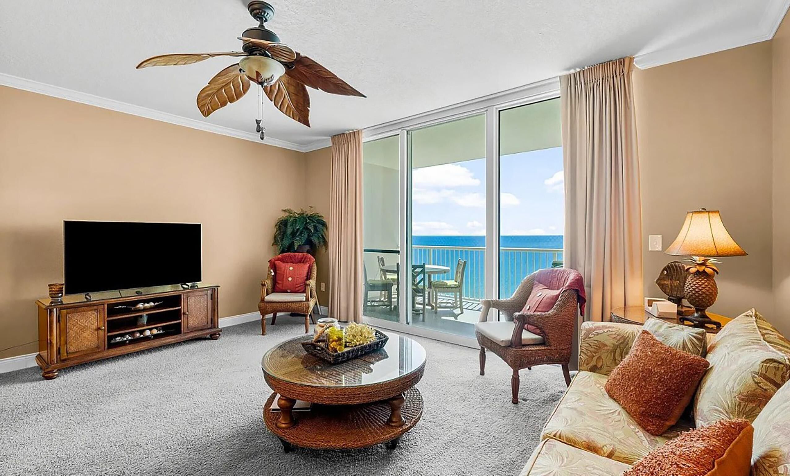 A living room with a flat-screen TV, wicker furniture, a ceiling fan, and a balcony with a view of the ocean.