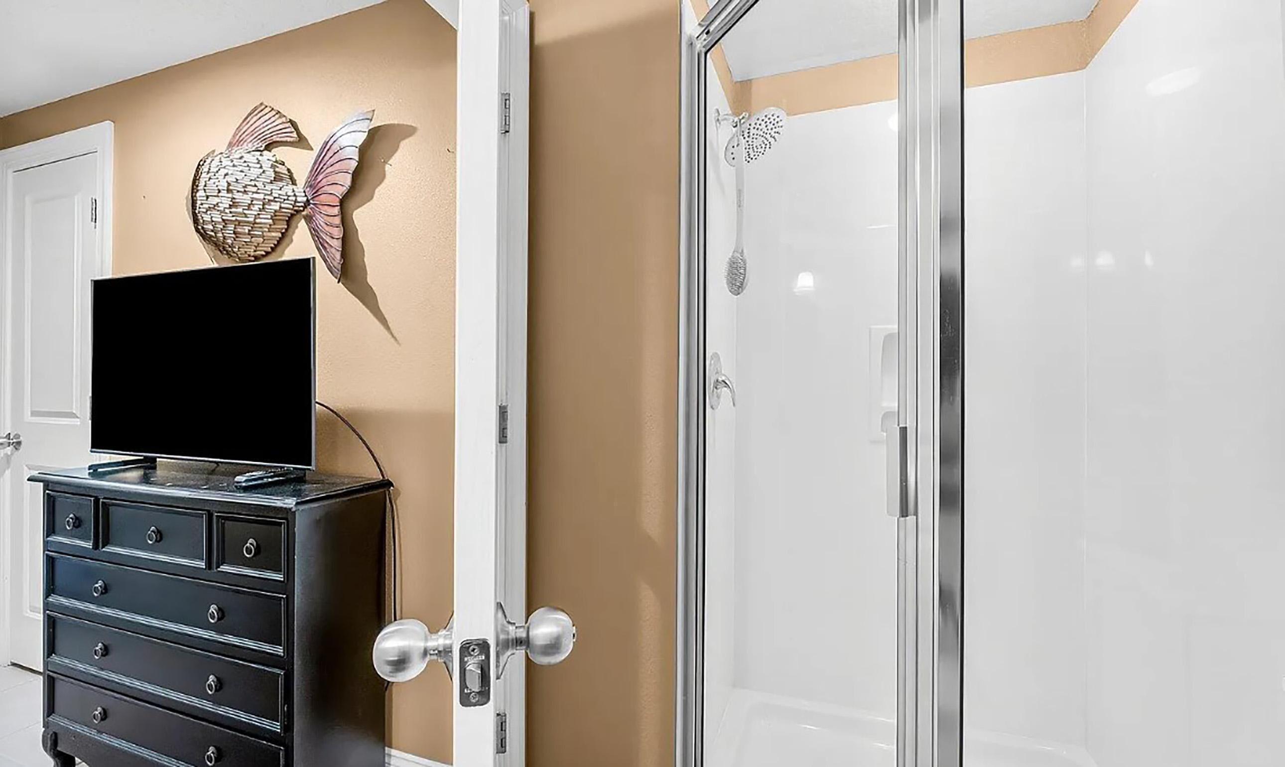 A hotel room with a TV on a dresser, a decorative wall piece, and a shower with a glass door.