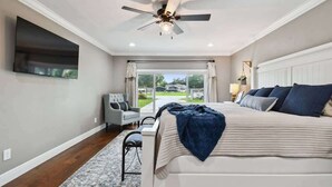 Ein Schlafzimmer mit einem großen Bett, einem Deckenventilator, einem an der Wand montierten Fernseher und Blick ins Freie durch eine Schiebetür.