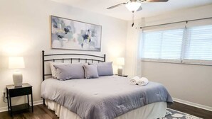 Une chambre à coucher comprenant un lit, deux tables de chevet, un ventilateur de plafond et une fenêtre avec des stores.
