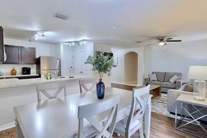 Eine moderne Küche mit weißen Schränken, ein Essbereich mit weißem Tisch und Stühlen und ein Wohnzimmer mit grauem Sofa und einer Lampe.