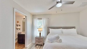 Quarto de hotel com cama, mesa de cabeceira, ventoinha no teto e banheiro visível por uma porta aberta.