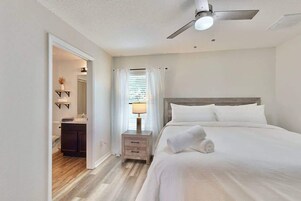 Habitación de hotel con cama, mesita de noche, ventilador de techo y baño visible a través de una puerta abierta.
