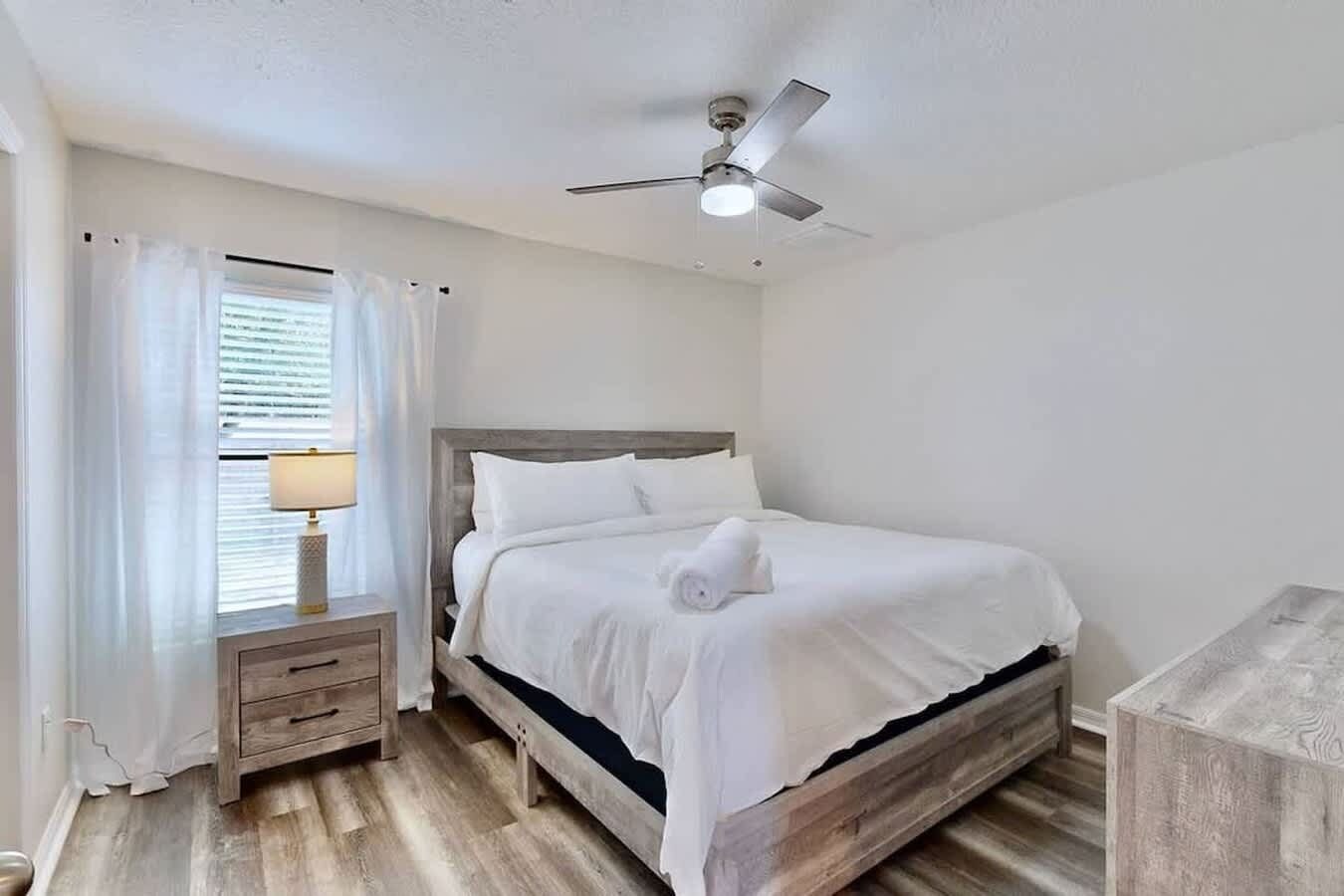 A bedroom with a bed, a nightstand, a lamp, a ceiling fan, and a window with curtains.