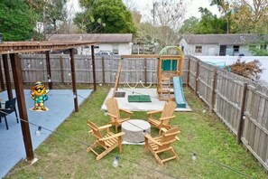 Ein Garten mit einer Holzterrasse, einer Schaukel, einer Rutsche und einem Tisch mit Stühlen.