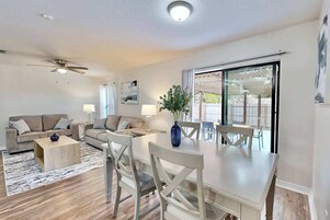 Un salón espacioso con comedor, ventilador de techo y un amplio ventanal corredera que da a un patio exterior.