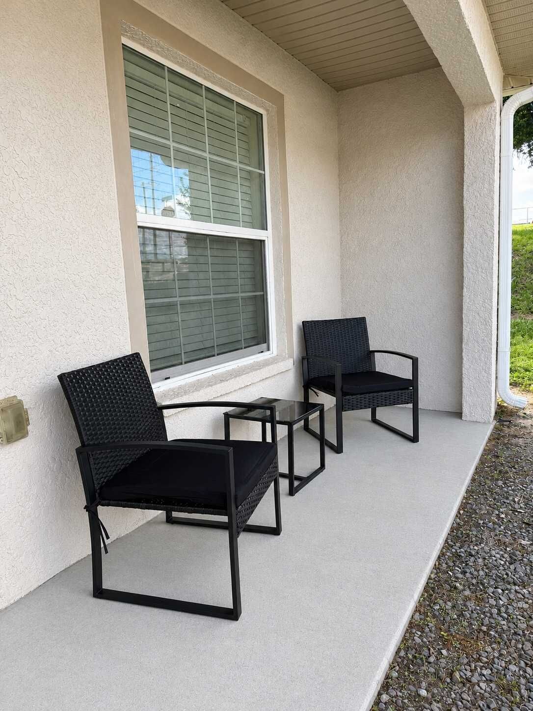 Un espacio de estar al aire libre cubierto con dos sillas y una mesa pequeña, junto a una ventana con persianas.