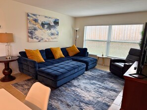 A living room with a blue sofa, a small round table, a lamp, and a large window with blinds. - Contemporary 3 Bedroom Home in Dade City (Dade City)